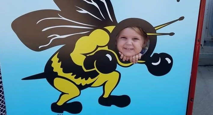 little girl looking through hole in board with mascot printed on it (yellow jacket)