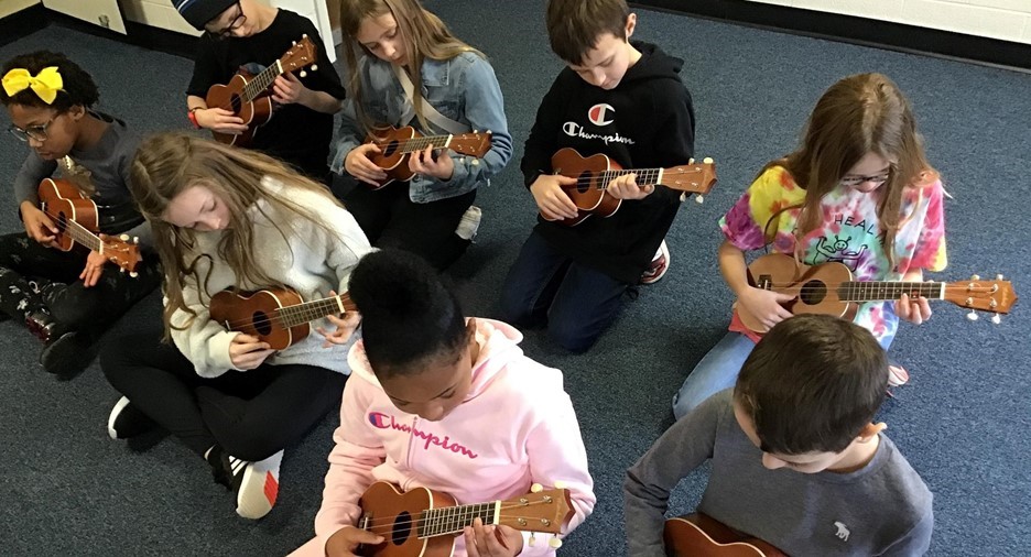 8 students sitting on the floor playing ukuleles